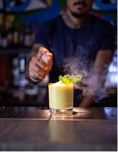 Mint & Mango Cocktail im Old Fashioned Glas, gelb, mit Minzgarnitur, Bartender im Hintergrund