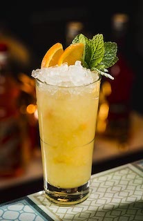 Nutty Mango Cobbler Cocktail im Highball Glas mit Crushed Ice, Orangenscheiben und Minze auf Bar