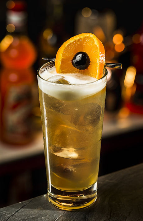 Orange Collins Cocktail im Highballglas mit Eis, Orangenscheibe und Cocktailkirsche, auf Bar-Theke bei warmem Licht serviert