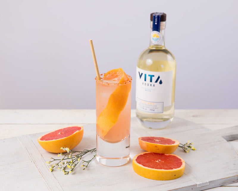 Palomita Cocktail mit Grapefruit und Eis im Highballglas, serviert neben Vodkaflasche und frischen Grapefruitstücken