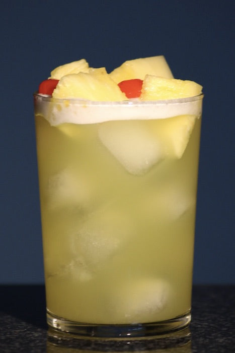 Pearl Harbor Cocktail im Highball-Glas auf Eis, mit Ananasstücken und roter Kirsche als Garnitur.