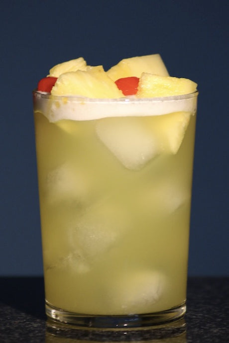 Pearl Harbor Cocktail im Highball-Glas auf Eis, mit Ananasstücken und roter Kirsche als Garnitur.