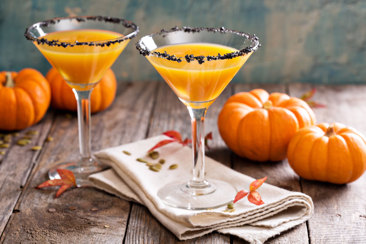 Zwei Pumpkin Margarita Cocktails mit schwarzem Salzrand auf Holztisch, dekoriert mit kleinen Kürbissen und Herbstblättern. 