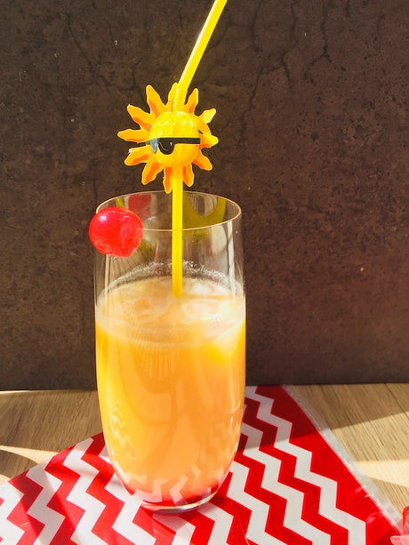 Red Sunshine Cocktail im Highballglas mit Eis, Kirsche und gelbem Strohhalm, serviert auf Tisch mit roter Serviette