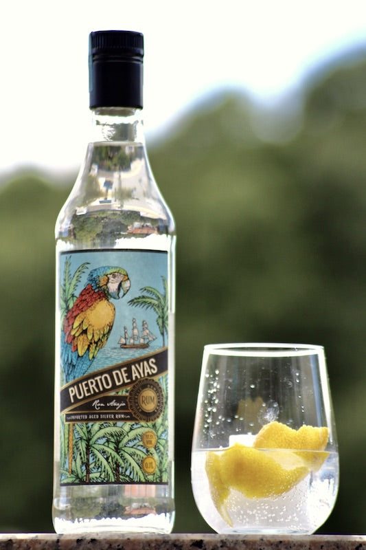 Puerto de Ayas Rum Flasche neben Glas mit Eis und Zitronenzeste vor unscharfem Hintergrund