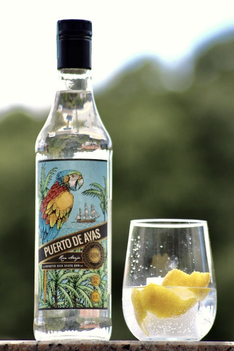 Puerto de Ayas Rum Flasche neben Glas mit Eis und Zitronenzeste vor unscharfem Hintergrund