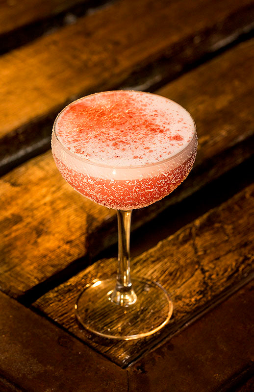 Rose Sunset Sour Cocktail im Coupette-Glas mit rosafarbener Schaumkrone und feinem Farbverlauf auf dunkler Holzoberfläche.