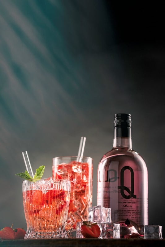 Rose Gin & Tonic mit Rose Gin Q Flasche, zwei Gläsern, Eis und Minze vor dunklem Hintergrund