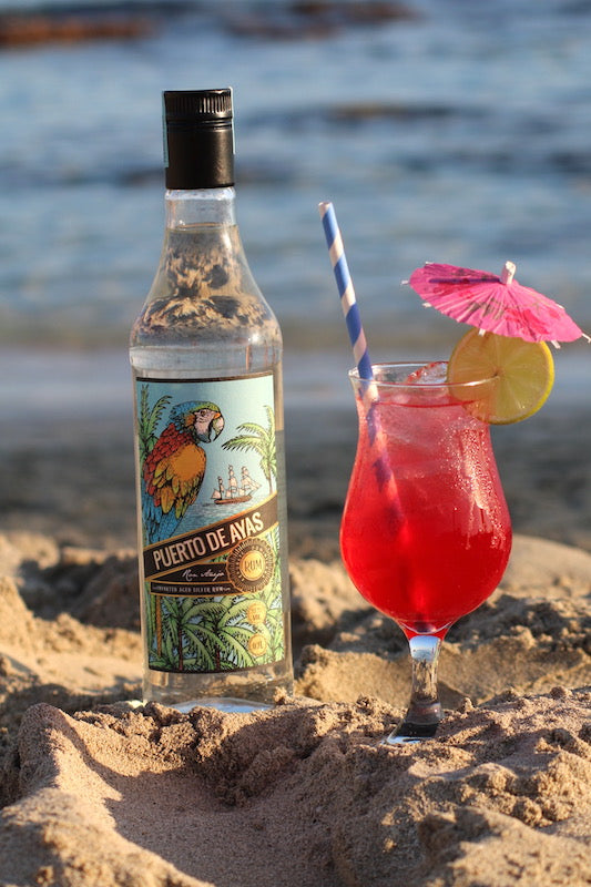 Rum Daisy Cocktail im Glas mit Eis, Zitronenscheibe und pinkem Schirmchen neben Rumflasche am Strand