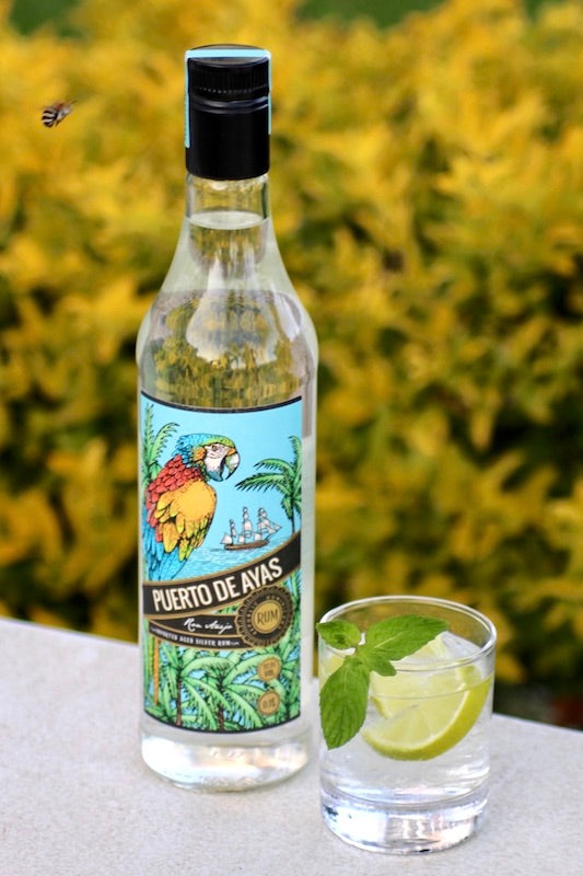 Flasche Puerto de Ayas Rum mit Cocktail im Glas, garniert mit Limettenscheibe und Minze, vor gelbem Hintergrund.