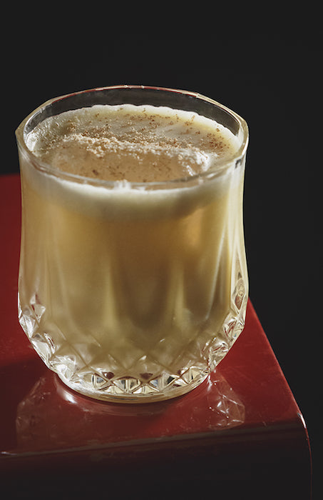 Rye Bite Cocktail in geschliffenem Tumbler-Glas mit cremigem Schaum und Muskat bestäubt auf einer roten glänzenden Oberfläche.