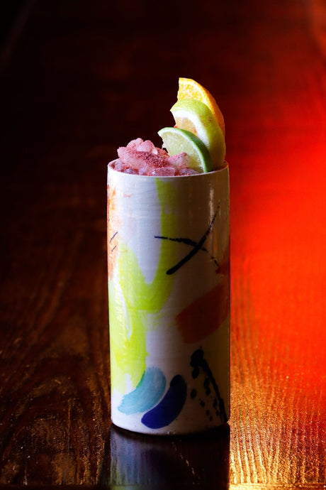 Farbenfroher Samba Cocktail mit Crushed Ice, Limette und Orange in bemaltem Keramikbecher auf dunkler Holzfläche.