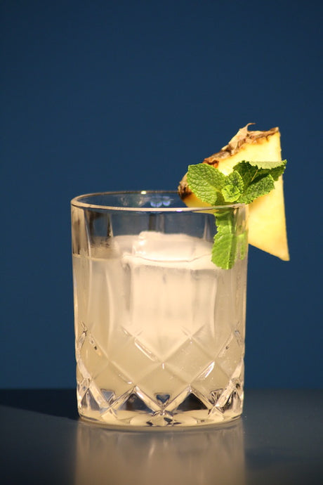 Saturn Cocktail in Kristallglas mit Ananasstück und Minze als Garnitur vor blauem Hintergrund.