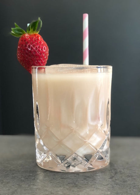 Cremiger Sombrero Mix Cocktail in einem Kristallglas mit Erdbeergarnitur und gestreiftem Strohhalm vor dunklem Hintergrund.
