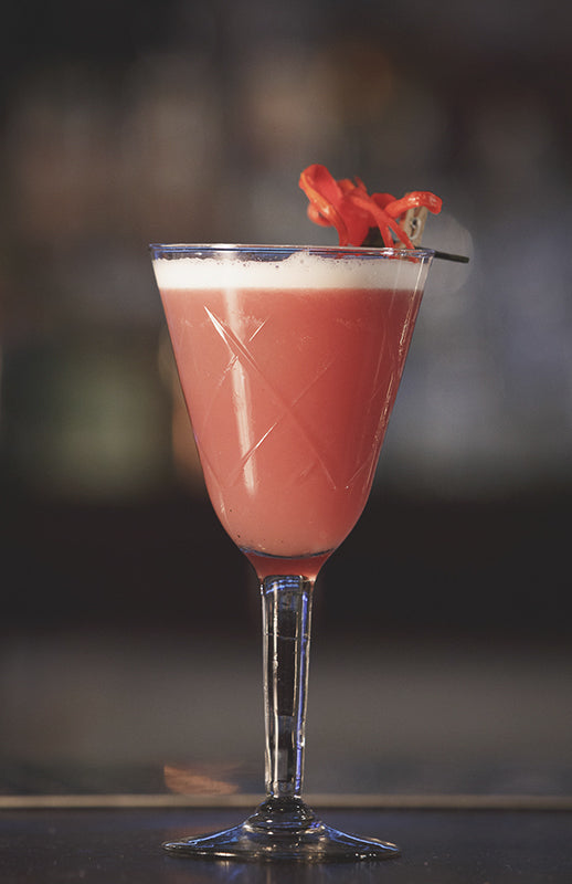 Sour Thai Jasley Cocktail im Cocktailglas mit Schaumschicht und roter Dekoration auf Bar vor unscharfem Hintergrund