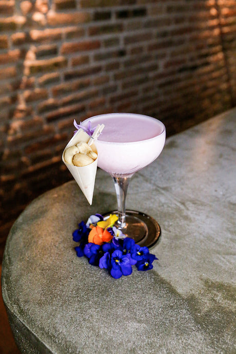 Strawberry Colada Fresón Cocktail in rosa Farbe mit Nuss-Tüte am Glasrand und violetten Blüten auf Betontisch.