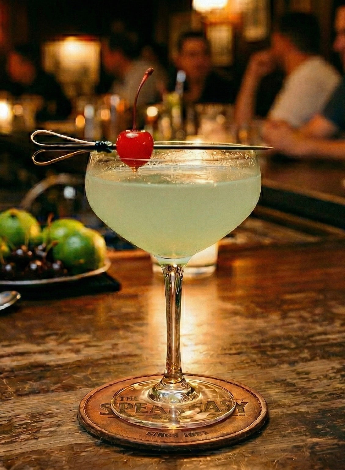 The Last Word Cocktail in Coupeglas mit Cocktailkirsche auf Holzbar, warme Bar-Atmosphäre im Hintergrund.