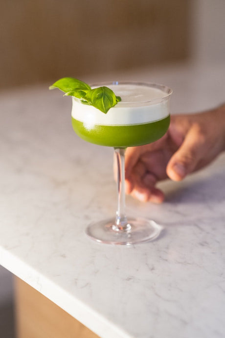 Grüner „Princess and the Pea“-Cocktail mit cremiger Schaumkrone, dekoriert mit Basilikumblatt, auf hellem Marmortisch.