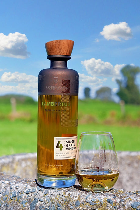 Belgischer Whisky Lambertus Single Grain Flasche mit Glas auf Steinmauer vor grünem Feld und blauem Himmel
