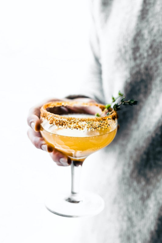 Thym'Punch Cocktail im Coupette-Glas mit Gewürzrand und Thymianzweig, von einer Person in der Hand gehalten