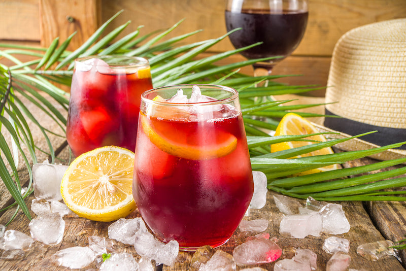 Tinto de Verano mit Rotwein, Zitronenscheibe und Eis im Glas auf Holztisch mit Palmblatt und Zitrone