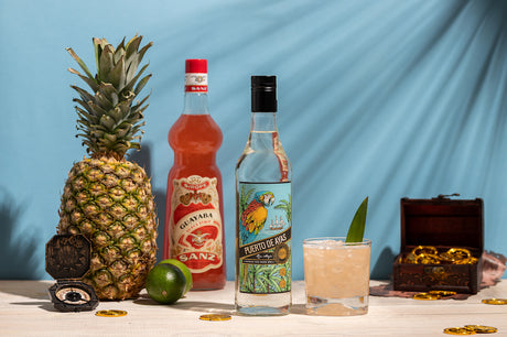 Tobago Cocktail mit Puerto de Ayas Rum, Guave-Sirup, Ananas, Limette, Kompass, Schatztruhe und Goldmünzen auf hellem Tisch