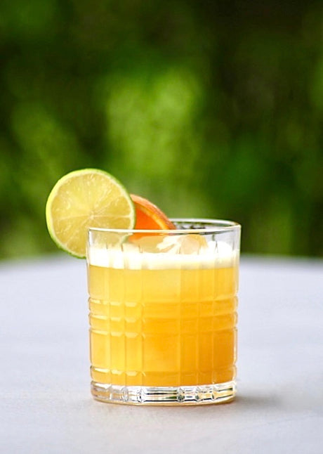 Alkoholfreier Tropical Fling Cocktail in einem Glas, dekoriert mit Limetten- und Orangenscheibe, serviert auf Eis vor einem grünen Hintergrund.