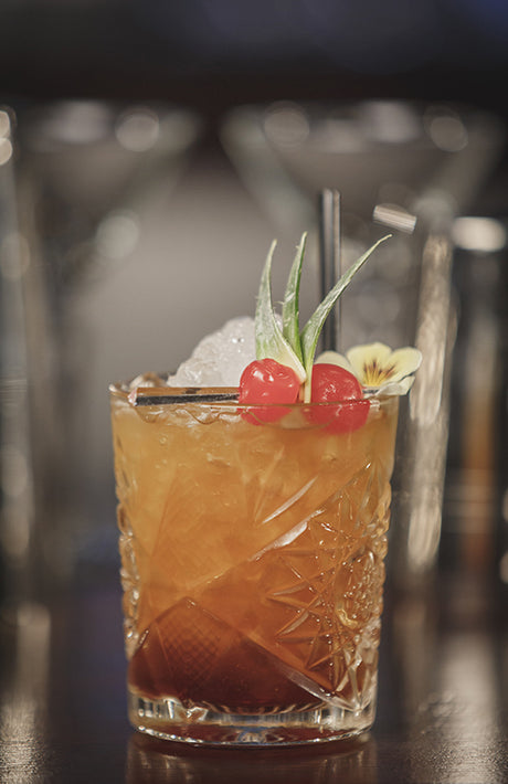 Tropical Fritz Cocktail im Tumbler mit Crushed Ice, Kirschen, Ananasblättern und Blüte dekoriert