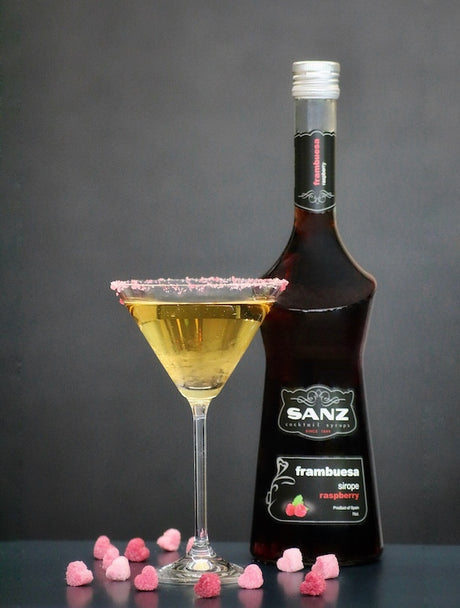 Vampire’s Kiss Cocktail im Martiniglas mit goldgelbem Getränk und rosa Zuckerrand, neben einer SANZ Himbeersirupflasche.