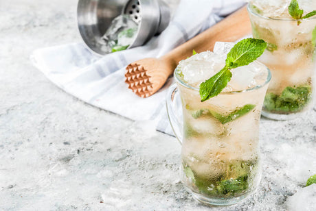Versinthe Mojito mit Minze und Crushed Ice im Glas, daneben Shaker und Barstößel auf heller Oberfläche