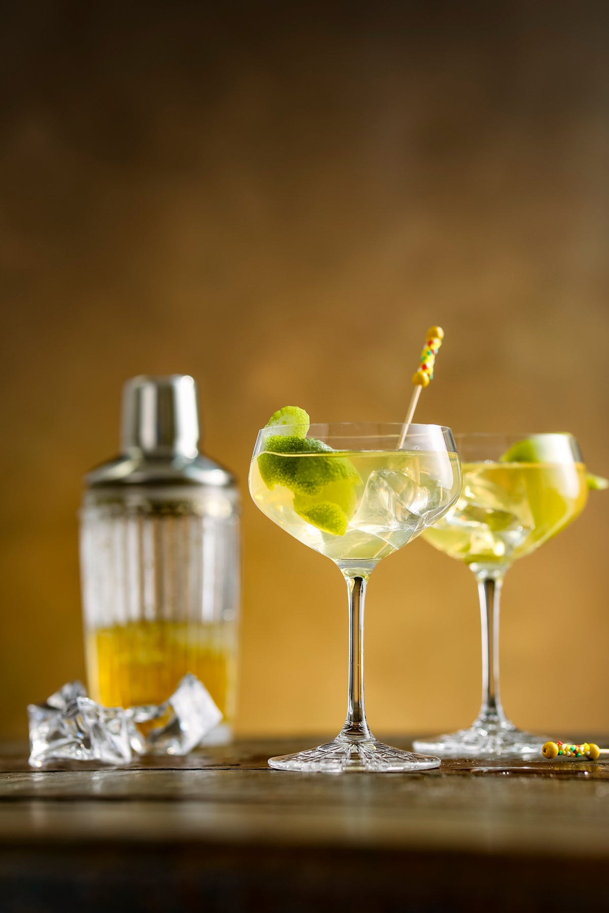 Zwei Yellow Submarine Cocktails in Coupette-Gläsern mit Limettenschalen, Shaker und Eiswürfeln im Hintergrund.
