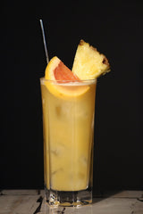 Acapulco Gold Cocktail im Highball Glas mit Eis, Ananasstück, Grapefruit und Trinkhalm vor dunklem Hintergrund