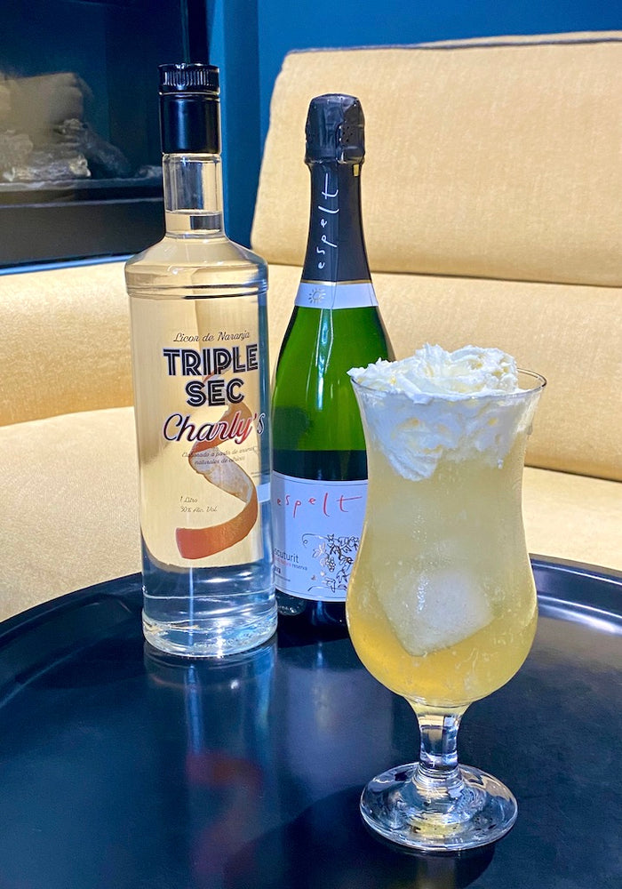 Agua de Sevilla Cocktail im Hurricane-Glas mit Cava und Triple Sec, Schlagobershaube, vor gelbem Sessel und blauem Hintergrund. 