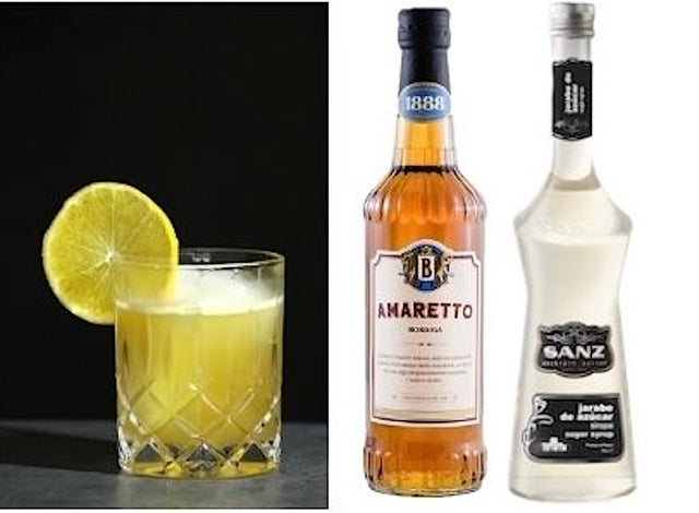 Amaretto Sour im Tumbler mit Zitronenscheibe, daneben Amaretto Bordiga und weißer Rohrzuckersirup von Sanz.