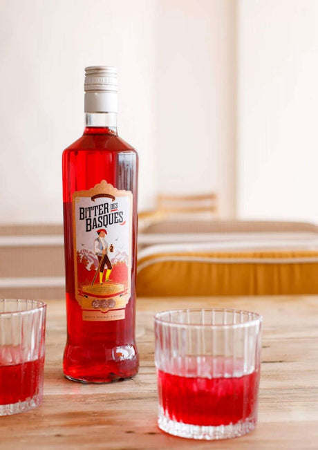 Bitter des Basques Flasche auf Holztisch mit zwei Gläsern, gefüllt mit rotem Aperitif, im Hintergrund gelbe Sitzpolster.