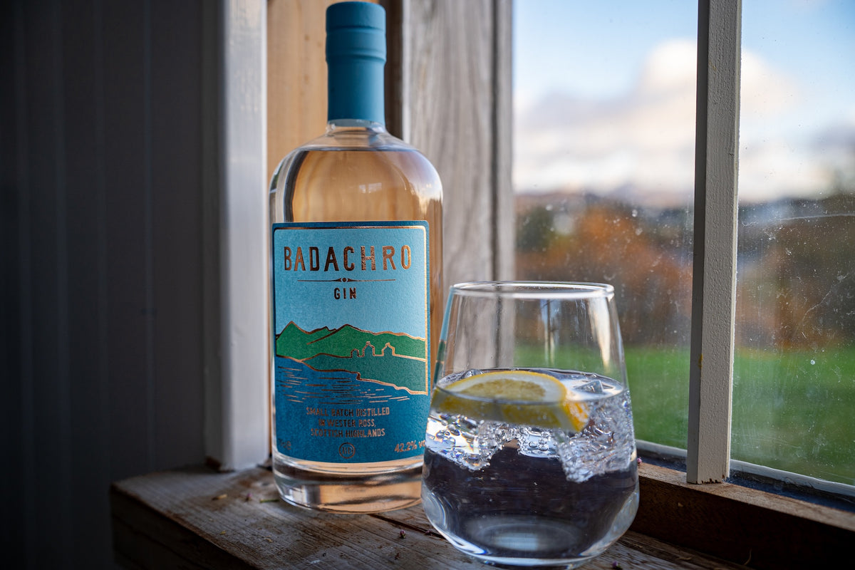 Flasche Badachro Storm Strength Gin neben einem Glas Gin & Tonic mit Zitronenscheibe auf einer Fensterbank mit Blick ins Grüne