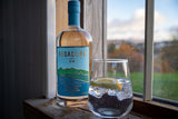 Flasche Badachro Storm Strength Gin neben einem Glas Gin & Tonic mit Zitronenscheibe auf einer Fensterbank mit Blick ins Grüne