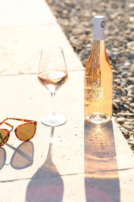 Rosé & Roll Weinflasche mit gefülltem Weinglas und Sonnenbrille auf heller Steinfläche, im Sonnenlicht mit Schattenspiel.