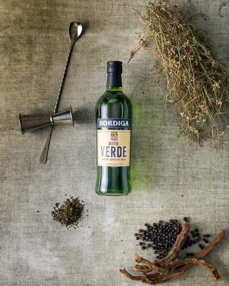Flasche Bordiga Bitter Verde mit Botanicals, Wacholderbeeren und Cocktailwerkzeugen auf grünem Hintergrund arrangiert.