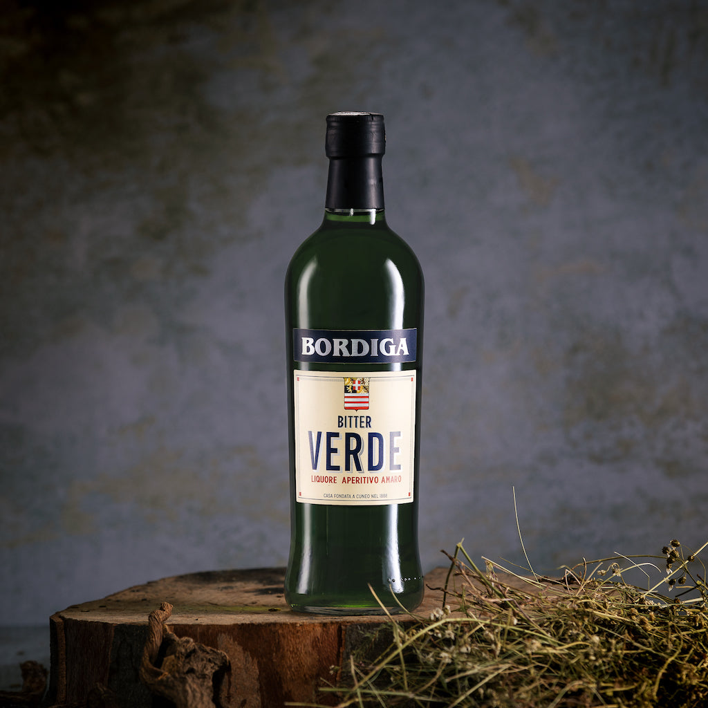 Flasche Bordiga Bitter Verde auf rustikalem Holzstumpf, umgeben von natürlichen Kräutern vor einem dunklen Hintergrund.