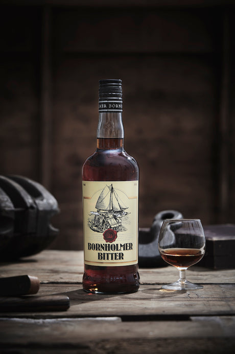 Bornholmer Bitter Flasche neben einem gefüllten Glas, präsentiert auf rustikalem Holztisch, stimmungsvolle Beleuchtung.