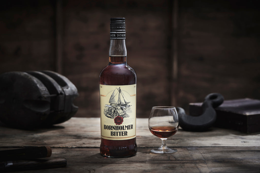 Bornholmer Bitter Flasche und Glas auf rustikalem Holztisch mit dunklem Hintergrund, umgeben von schattenhaften Gegenständen.