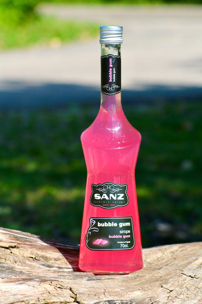 Bubble Gum Sirup Sanz 700 ml in einer pinkfarbenen Flasche mit schwarzem Etikett, auf einem Holzstamm platziert.