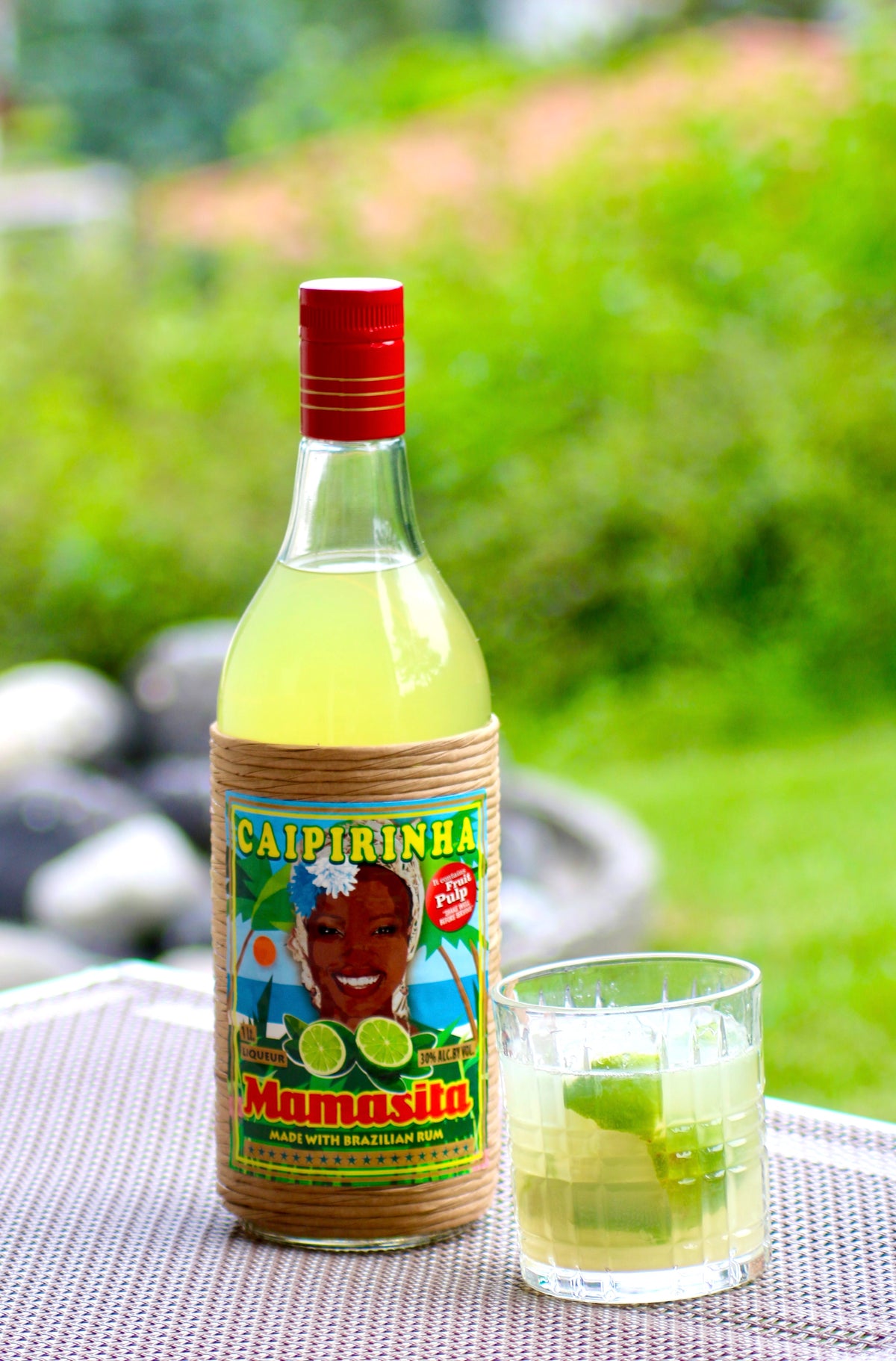 Mamasita Caipirinha Mix Flasche mit Caipirinha Glas und Limette auf Tisch vor grünem Hintergrund