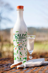 Flasche Calin Polisson Likör mit floraler Gestaltung, daneben Likörglas und Calissons auf Gartentisch vor Naturhintergrund.