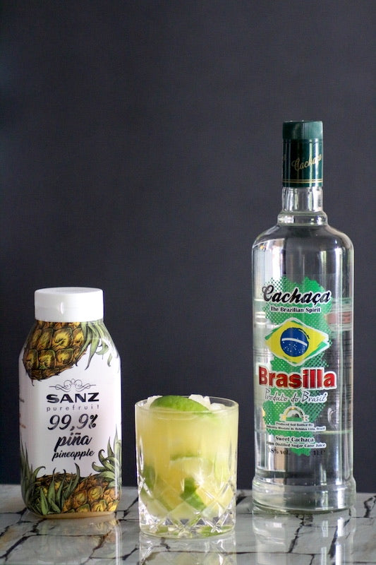 Ananas Caipirinha im Glas mit Limette und Eis neben Cachaça Flasche und Ananaspüree auf Tisch