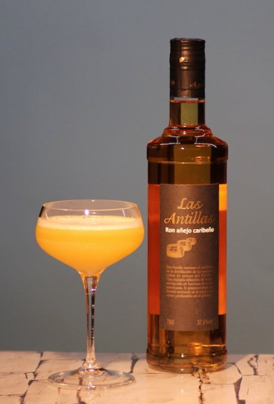Chinese Itch Cocktail in Orange mit feiner Schaumkrone in einer Cocktailschale neben einer Flasche Las Antillas Rum.