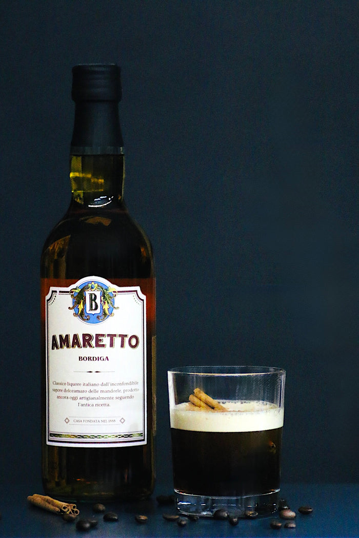 Amaretto Bordiga-Flasche neben dem Coffee Shadow Cocktail im Rocks-Glas, dekoriert mit Zimtrinde und Kaffeebohnen.