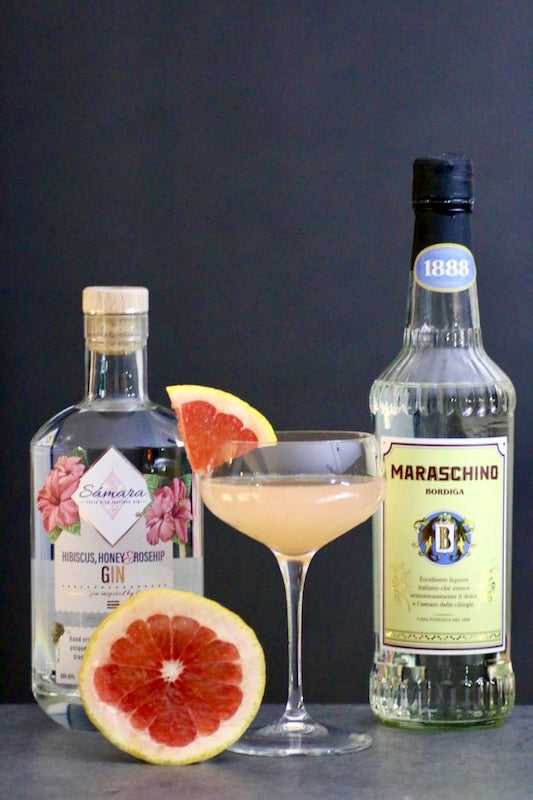 Colonial Cocktail mit Gin und Maraschino im Glas, garniert mit Grapefruit, daneben Flaschen und halbe Grapefruit