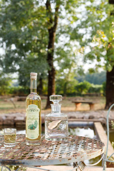 Flasche Fleur d'Angélique Likör auf einem Metalltisch mit Glas und Karaffe, im Hintergrund Bäume und ein Teich.